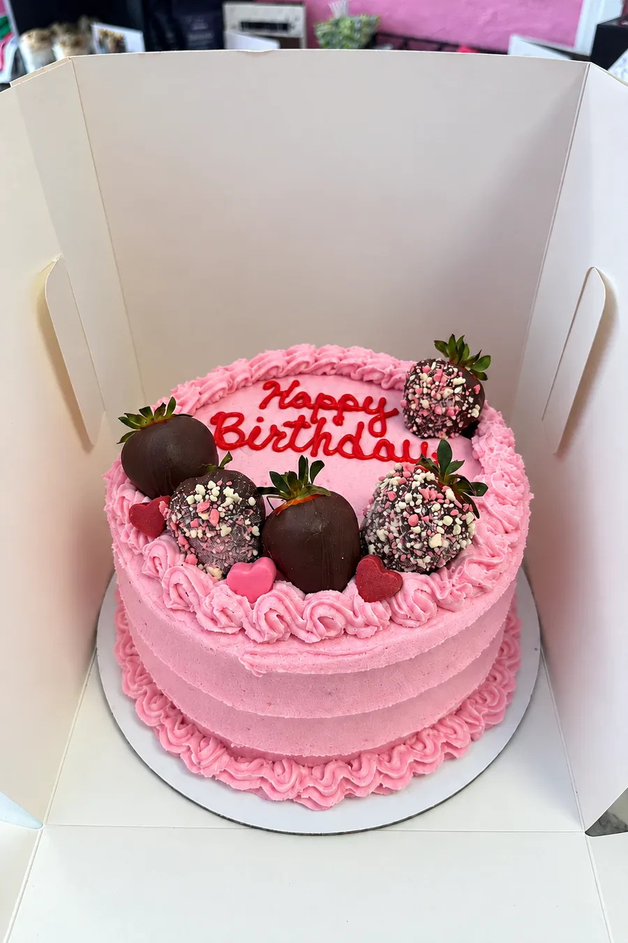 Pink Happy Birthday cake topped with chocolate covered strawberries, in an open bakery box